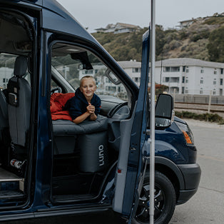 A child is lying inside a blue camper van, resting on a bed with a red sleeping bag. The van's side door is open, and an apartment building is visible in the background. The child is smiling and appears relaxed.