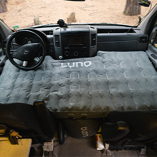 Interior view of a vehicle showing the dashboard and front seats. There is a large, inflated mattress laid over the seats and dashboard, extending from one side to the other. The logo "Luno" is visible on the mattress. The outdoors is faintly visible through the windshield.
