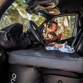 A child wearing a leopard-print outfit sits playfully on the passenger seat of a vehicle, with a forest visible outside the window. The vehicle has a mattress spread across the front seats, and a bright, sunny day is hinted at through the trees.