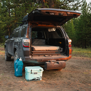 An SUV is parked with its rear hatch open, revealing a bed setup inside the vehicle. On the ground behind it, there is a blue water container and a light blue cooler. The scene is set in a forested area during daylight.