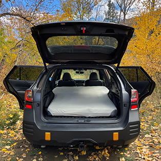 A car with its back hatch open reveals a neatly made bed in the cargo area. The car is parked outdoors, surrounded by trees with yellow and orange autumn leaves. The left and right rear doors are also open, providing a full view of the cozy setup.