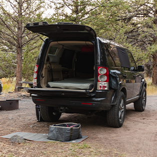 A black SUV is parked in a forested area with its rear hatch open, revealing a sleeping area inside. The ground around the vehicle has a mat and some camping gear. Tall trees are visible in the background.