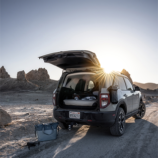 A parked SUV with its tailgate open faces a desert landscape during sunset. The sun peeks through the vehicle, casting long shadows. Camping gear is visible inside the SUV, and a cooler and tripod are placed on the ground next to it. Rocky formations are in the background.