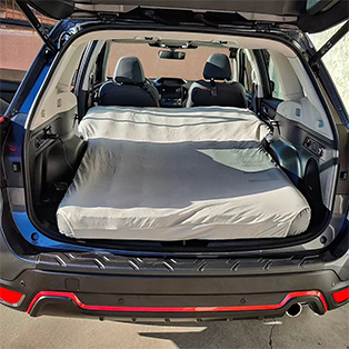 An SUV with its rear hatch open, revealing a neatly made mattress placed in the cargo area, creating a temporary sleeping setup. The front seats are visible, and sunlight illuminates the back of the vehicle.
