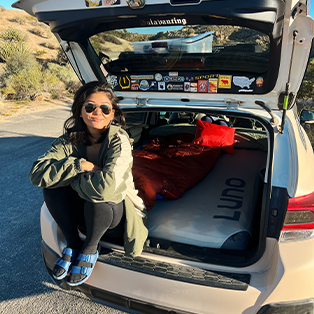 A person wearing sunglasses and casual clothes sits on the edge of an open car hatchback, which is set up with a mattress and bedding in the back. The car is parked on a sunny day by a desert landscape. The hatch door is adorned with various stickers.