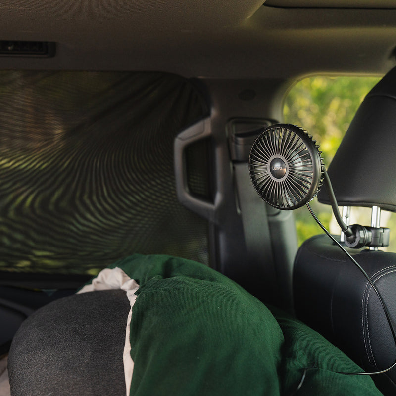 A Luno® Car Camping Cool Down Bundle, featuring a compact black fan clipped to the headrest and window screens, is set up in a car with a green blanket on the seat and sunlight streaming through the screened window.
