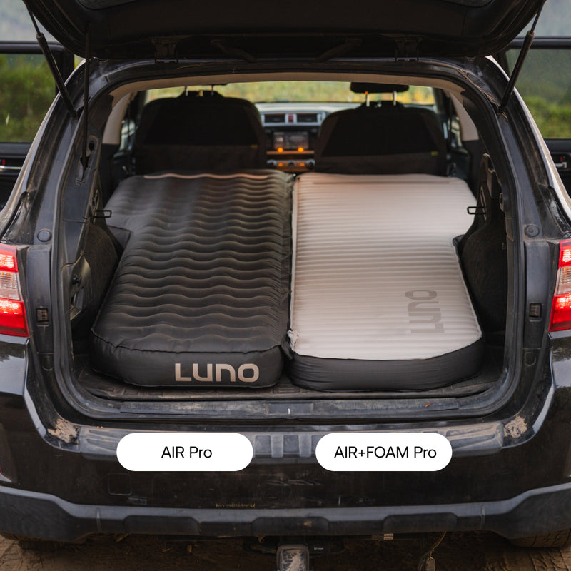 A Subaru Outback trunk with back seats down holds two Subaru Outback Car Camping Mattresses: the left one, labeled AIR Pro, is black; the right, labeled AIR+FOAM Pro, is white and black.