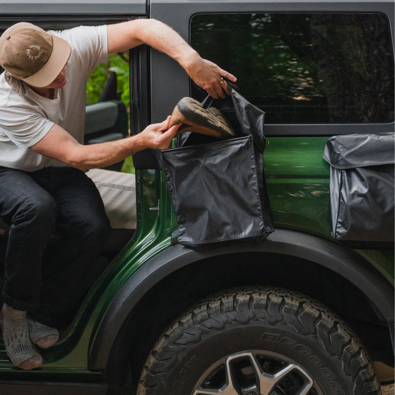 Wearing a tan hat, white shirt, and dark pants, a person in a green off-road vehicle organizes shoes and gear into the water-resistant Luno® Gear & Shoe Storage Bag attached to the rear side, keeping essentials sorted and accessible.