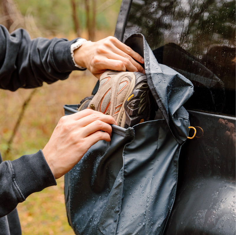 A person stores hiking shoes in the Luno® Gear & Shoe Storage Bag, attached to a vehicle’s back. The water-resistant fabric features visible raindrops, with lush greenery in the background.