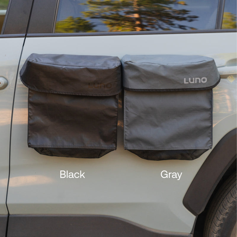 Two water-resistant Luno® Gear & Shoe Storage Bags, one black and one gray, are attached to a light-colored vehicle door. Each bag features a flap closure, bottom mesh pocket, and doubles as a gear pouch.