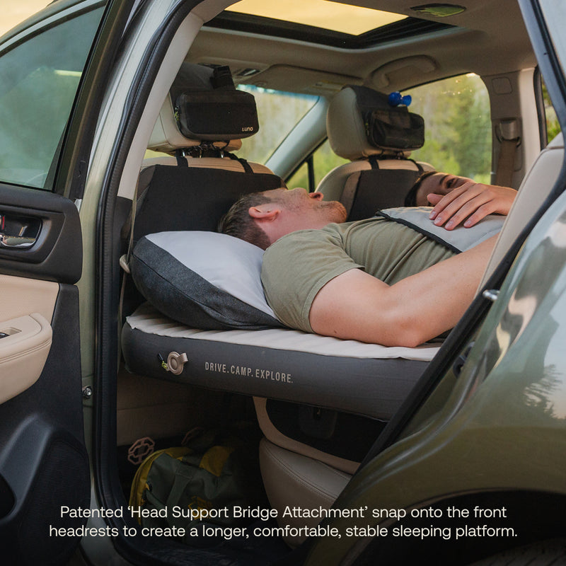A man relaxes on the AIR+FOAM Pro Car Camping Mattress in a car's backseat with the rear door open. The mattress spans the seats, providing a stable, comfy sleeping platform ideal for camping or road trips.