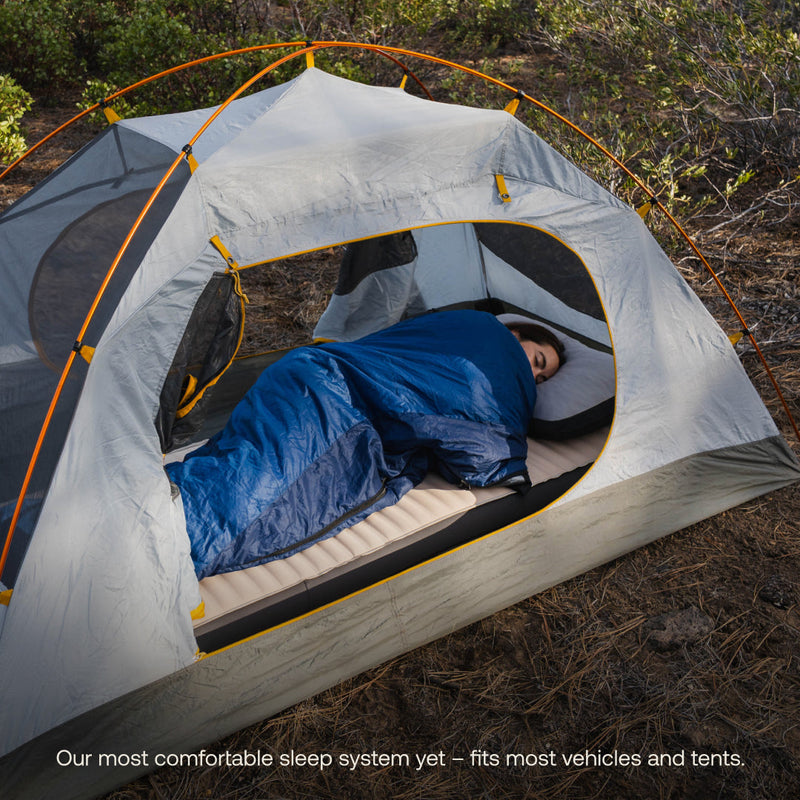 A person is comfortably sleeping on a pre-owned AIR+FOAM PRO Camping Mattress by Luno HQ, in like-new condition, inside a gray and yellow tent set up in a forested area. They are covered with a blue sleeping bag. Text at the bottom reads, "Our most comfortable sleep system yet – fits most vehicles and tents.