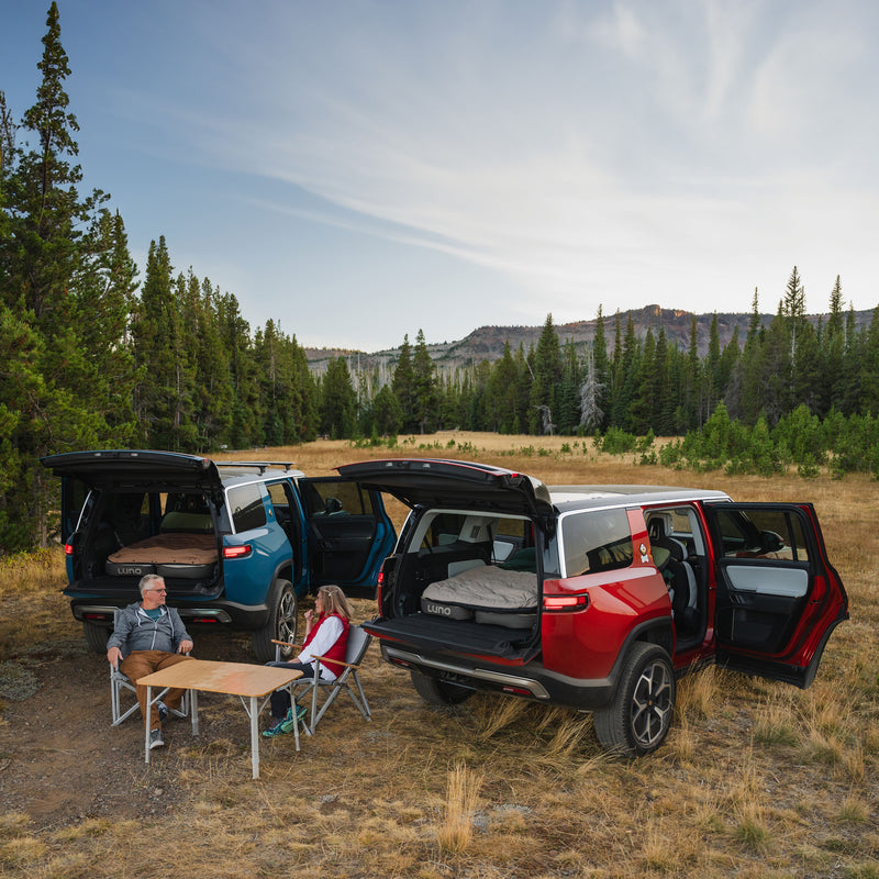 Two people relax at a folding table and chairs near two parked SUVs with open trunks, set up for camping. In one trunk, the AIR+FOAM Pro - Rivian R1S Compatible sleep system offers comfort amid pine trees and distant hills.