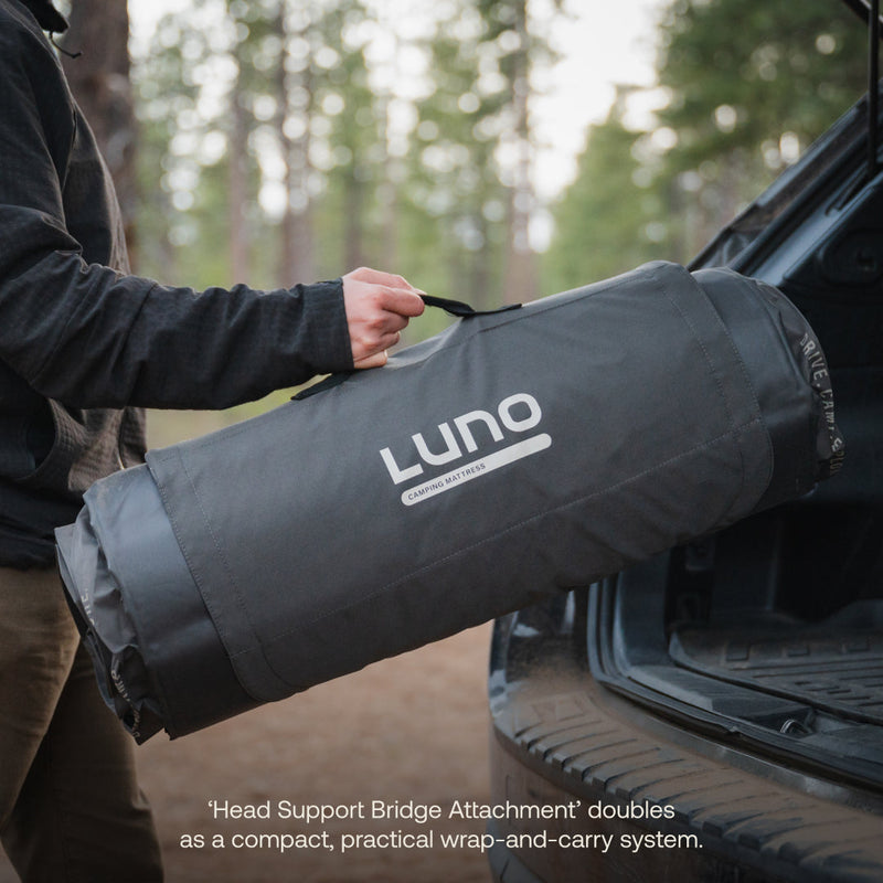 A person places a rolled-up AIR+FOAM PRO Camping Mattress by Luno HQ, in pre-owned but like-new condition, into the trunk of a car. This cylindrical mattress features a carrying handle, with trees and hints of outdoor scenery in the background.