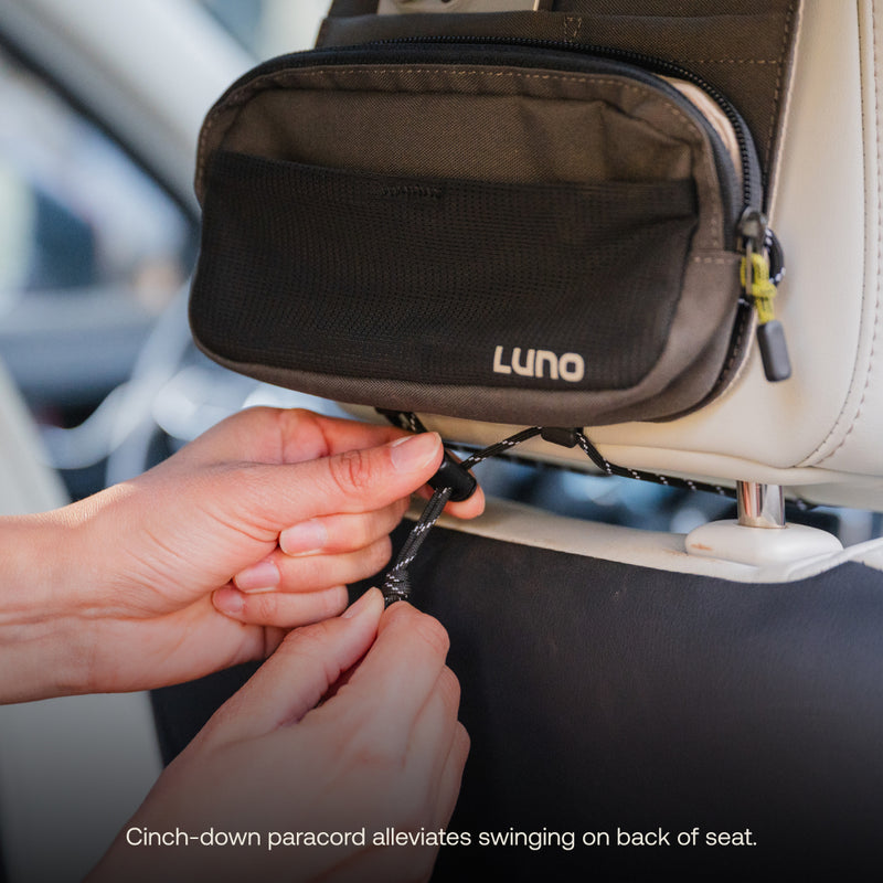 Hands are securing a pre-owned Luno HQ Headrest Organizer in black to the back of a car seat using cinch-down paracord, providing a practical and stable solution. The easy-to-install design ensures it stays in place while driving. Text at the bottom reads, "Cinch-down paracord alleviates swinging on back of seat.