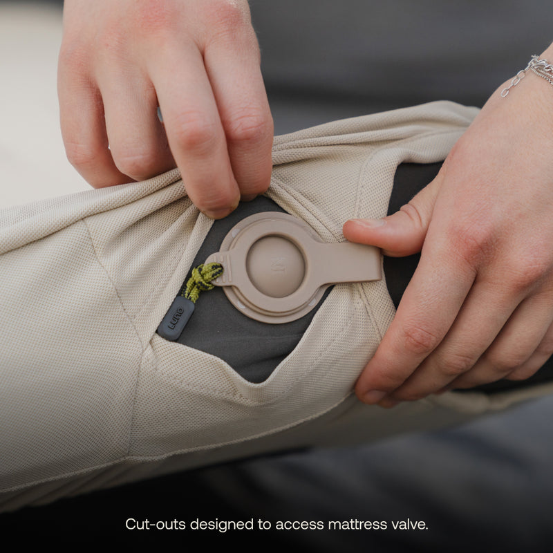 Close-up of a person’s hands opening a small flap on a beige fabric item to access a round, grey mattress valve underneath. There is a small tag with "Luno®" written on it near the flap. The image text reads, "Fitted Mattress Sheet - AIR+FOAM Pro Camping Mattress designed with cut-outs to access mattress valve.