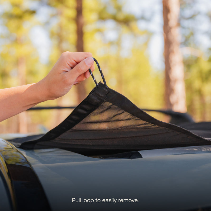 A hand tugging on a loop of the black mesh Sunroof Window Screen (Pre-Owned) from Luno HQ, displayed on a car roof for excellent airflow and insect protection, with blurred trees behind. Text says, "Pull loop to easily remove.