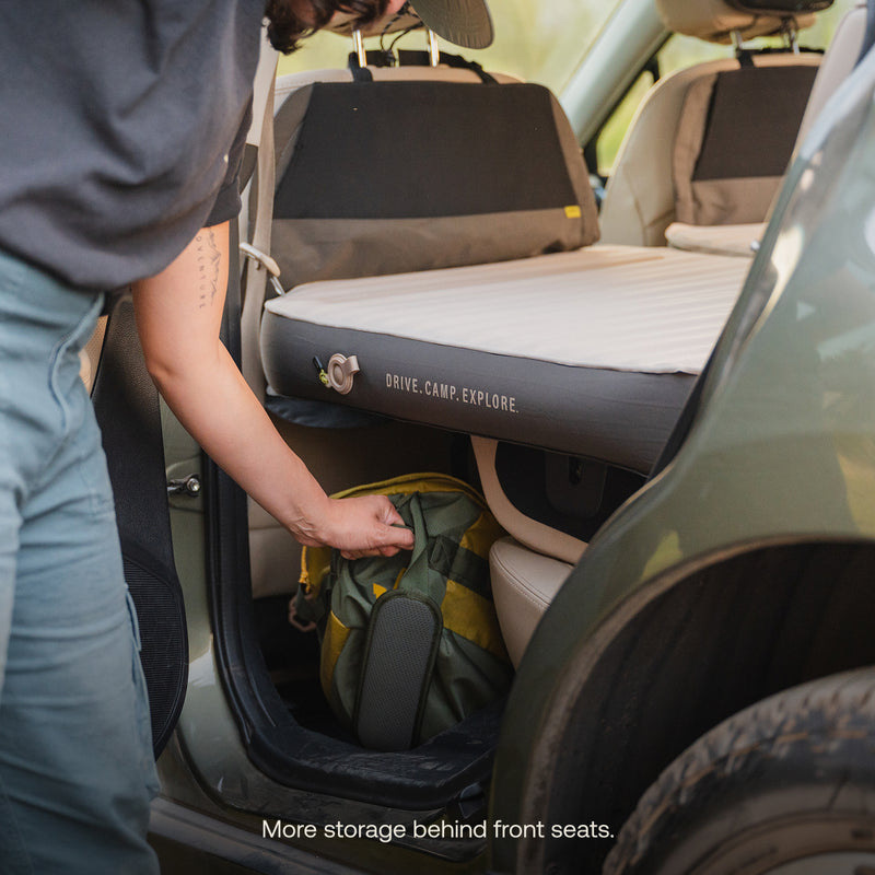 A person places a green and yellow backpack behind the front seats of a car with an AIR+FOAM Pro Car Camping Mattress set up in the back. Text on image: More storage behind front seats.