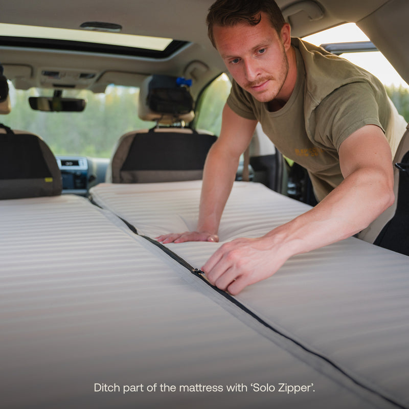 A man in a car unzips the middle of the AIR+FOAM Pro Car Camping Mattress laid across the back seats. Text reads: Remove part of the mattress with ‘Solo Zipper’.