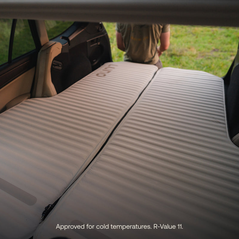 Two AIR+FOAM Pro Car Camping Mattresses are laid out side by side in a car, creating a sleeping surface. A person sits facing away outside the car with grass visible. Text: Approved for cold temperatures. R-Value 11.