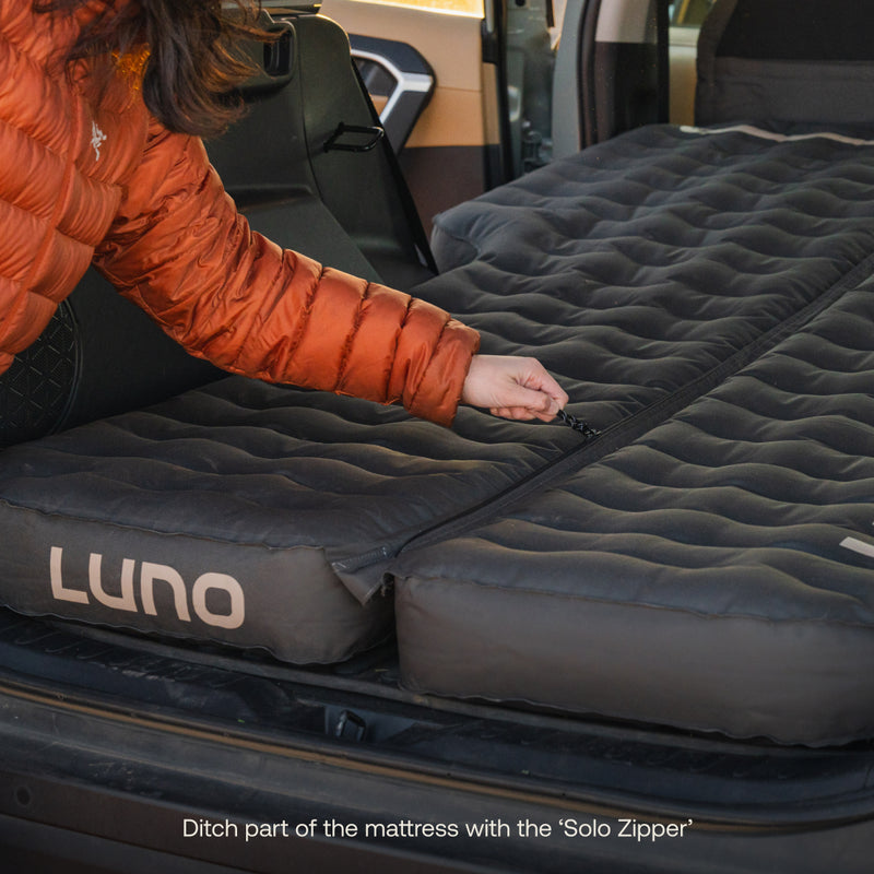 A person in an orange jacket unzips a Luno® AIR Pro Vehicle Mattress in the back of a vehicle, displaying the "Solo Zipper" for customizable flexibility.