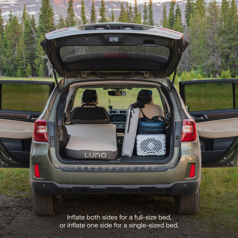 SUV with back hatch open displays an AIR+FOAM Pro Car Camping Mattress set up on one side, camping gear organized beside it. Forest and mountains are in the background. Bottom text highlights inflation options for the mattress.