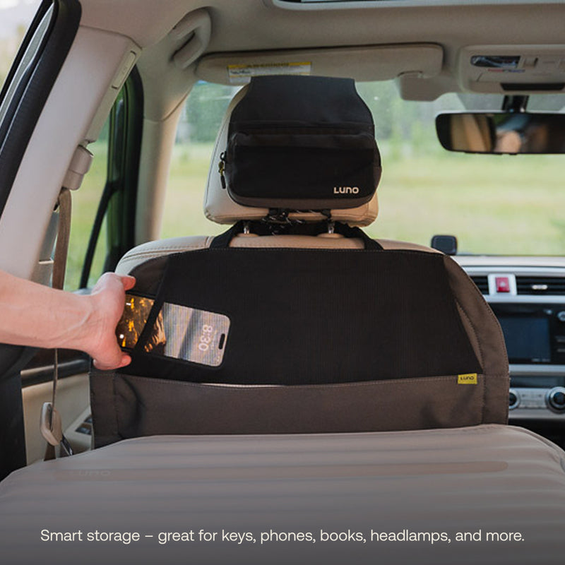 A person places a smartphone into a black and gray organizer attached to the back of a seat near the AIR+FOAM Pro Car Camping Mattress. An extra storage pouch is strapped to the headrest in a light-colored car interior.