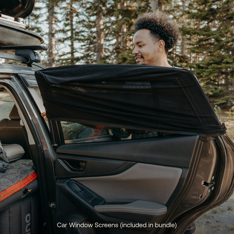 A person installs a mesh screen onto a car window as text overlay reads "Car Window Screens (included in the Car Camping AIR+FOAM Pro Starter Bundle)." The vehicle is parked in a forested area with the back door open, showing some camping gear inside. The perfect addition to your Luno® Car Camping adventure!