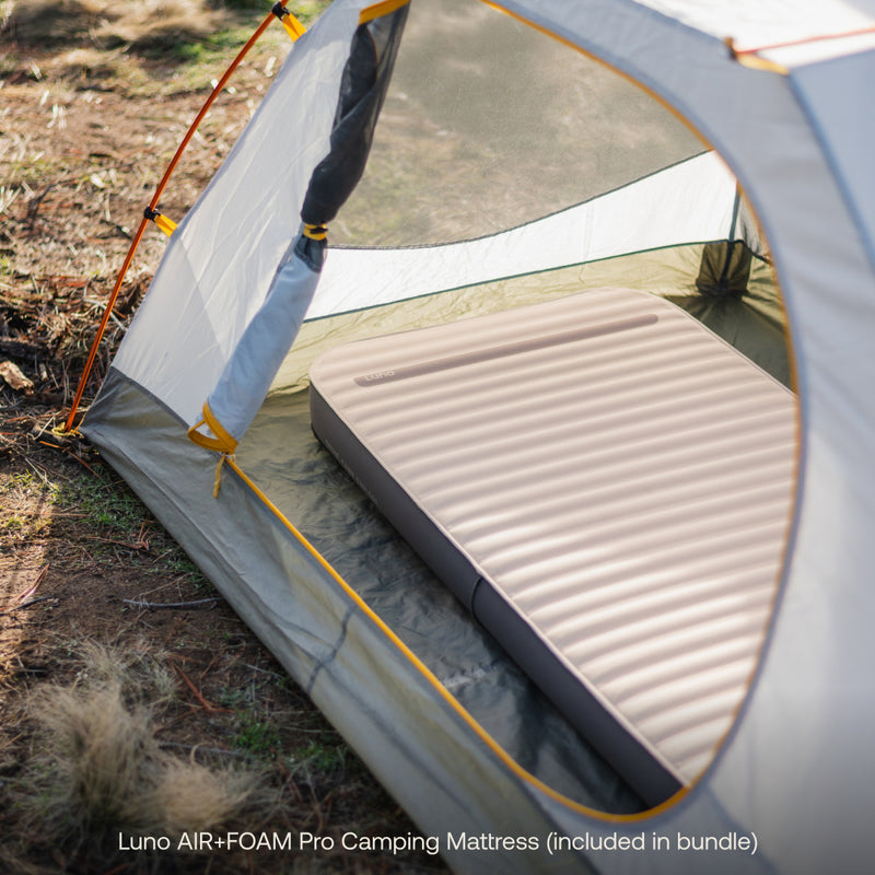 A tent with mesh panels and orange accents features an inflatable mattress, ideal for car camping. Text highlights "Luno® Camping AIR+FOAM Pro Starter Bundle," showcasing the ultimate Luno experience on dry grass.
