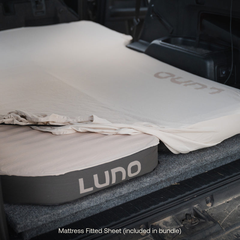 A mattress with a fitted sheet is set up in the back of a vehicle, perfect for those who enjoy Luno Car Camping. The mattress features the brand name "Luno®" printed on the side. The text at the bottom of the image reads, "Mattress Fitted Sheet (included in Car Camping AIR+FOAM Pro Starter Bundle).