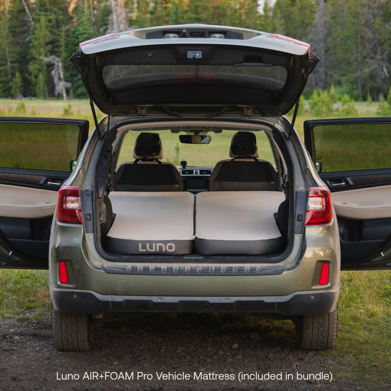 A parked SUV with its rear hatch open reveals a fully inflated Luno® Car Camping AIR+FOAM Pro Starter Bundle set up in the cargo space. The vehicle's back seats are folded down, creating a cozy haven perfect for car camping essentials amidst a grassy area and tall trees in the background.