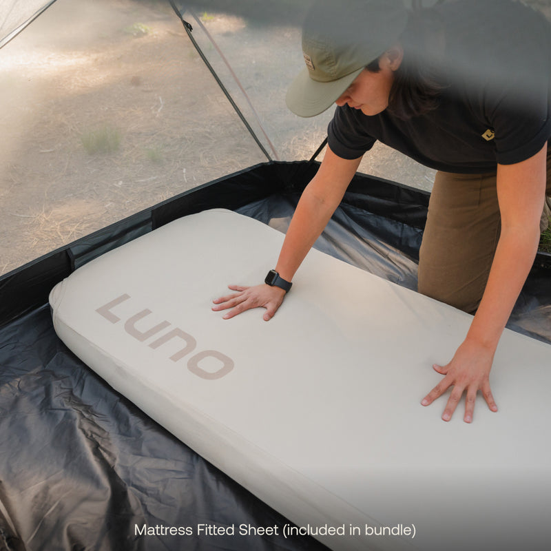 In the tent, someone adjusts the Luno® fitted sheet on an inflatable mattress—ideal for car camping. They wear a cap and smartwatch. Text at the bottom notes that this AIR+FOAM mattress fitted sheet is part of the Camping AIR+FOAM Pro Starter Bundle by Luno®.