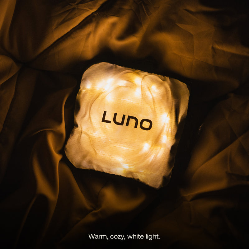 A glowing, square-shaped cushion with "Luno®" printed on it is illuminated by soft, warm white Luno® Camping String Lights. The cushion rests on a wrinkled, dark golden fabric background. Text at the bottom reads, "Warm, cozy, white light.