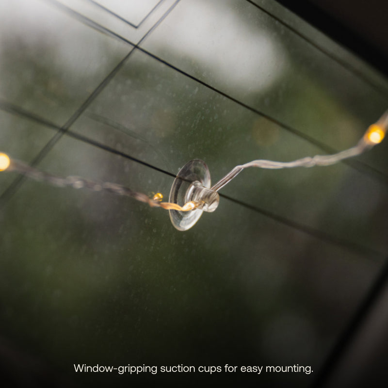 A close-up of Luno® Camping String Lights mounted on a window using a transparent suction cup. The background shows the blurred view through the window. The text below the image reads, "Window-gripping suction cups for easy mounting.