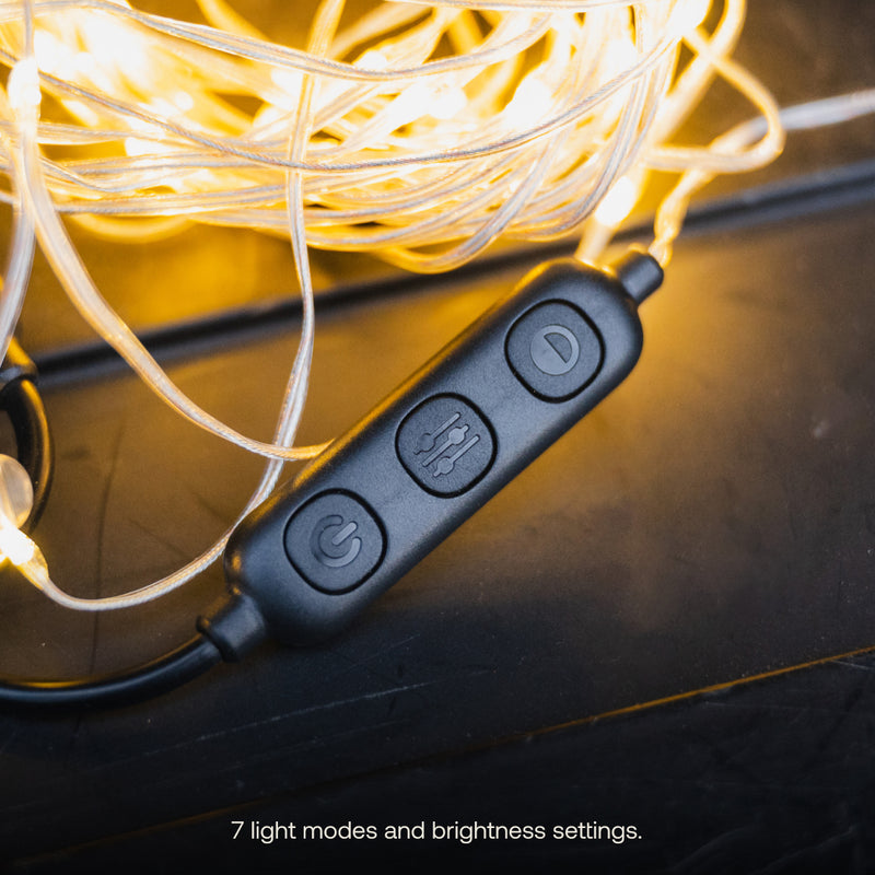 Close-up of a black remote control for Luno® Camping String Lights, with three buttons featuring icons: a power symbol, settings, and adjustable brightness. The remote is placed among glowing warm white LED camping string lights. Text at the bottom reads "7 light modes and brightness settings.