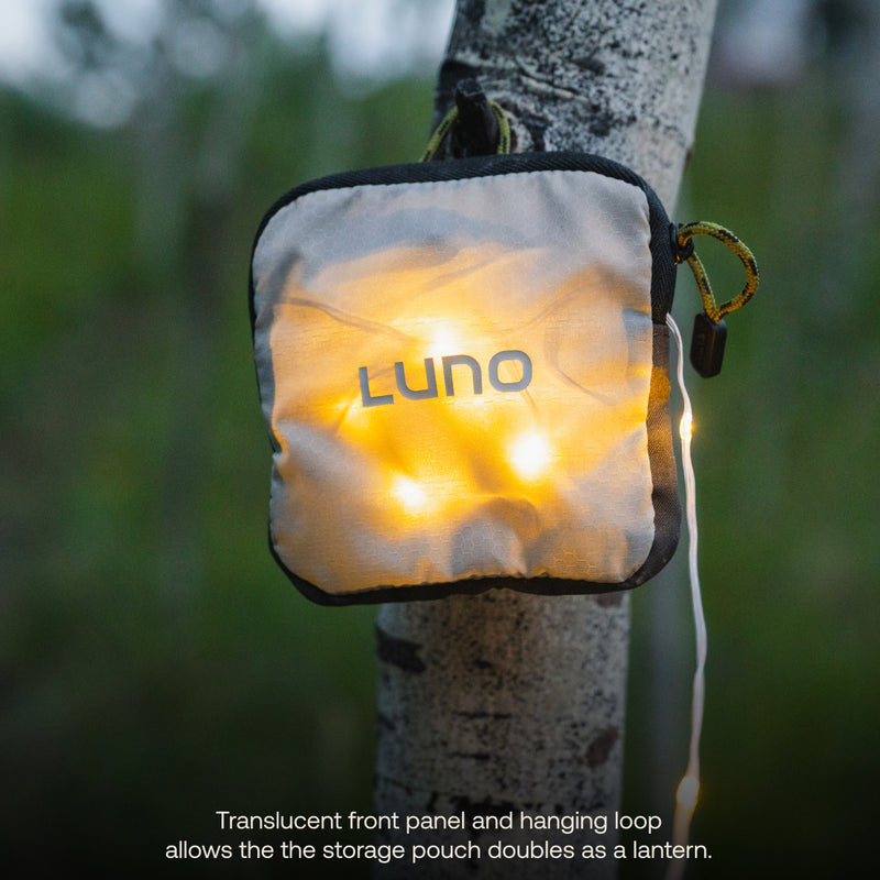 A soft, square-shaped storage pouch branded with "Luno®" gently illuminates with adjustable brightness, powered by a rechargeable USB-C battery pack inside. Hanging from a tree trunk using its loop, the pouch stands out against a blurred natural outdoor backdrop. Text at the bottom highlights its dual function as a lantern for the Camping String Lights product.