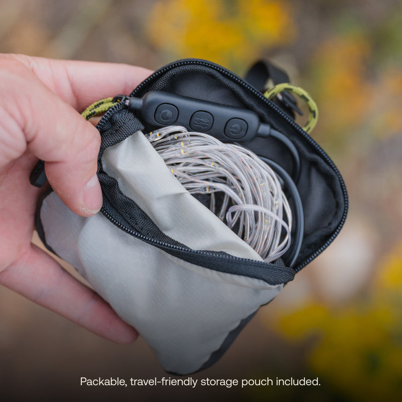 A hand is holding a compact, grey, and black zippered pouch that is partially open to reveal coiled earphones or wires inside. The text at the bottom of the image reads, "Packable travel-friendly storage pouch included." This versatile Luno® Camping String Lights pouch can also neatly fit your other small accessories for convenience.