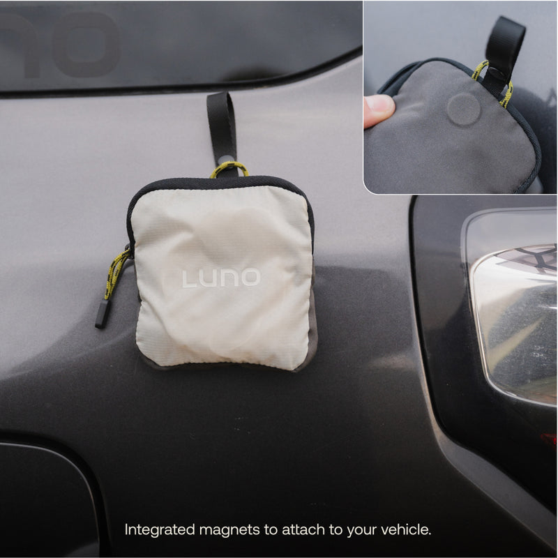 Small gray pouch labeled "Luno® Camping String Lights" attached to a car's exterior using integrated magnets. An inset image shows a close-up of the pouch’s corner, highlighting the magnet feature and its rechargeable USB-C battery pack. Text at the bottom reads: “Integrated magnets to attach to your vehicle.”
