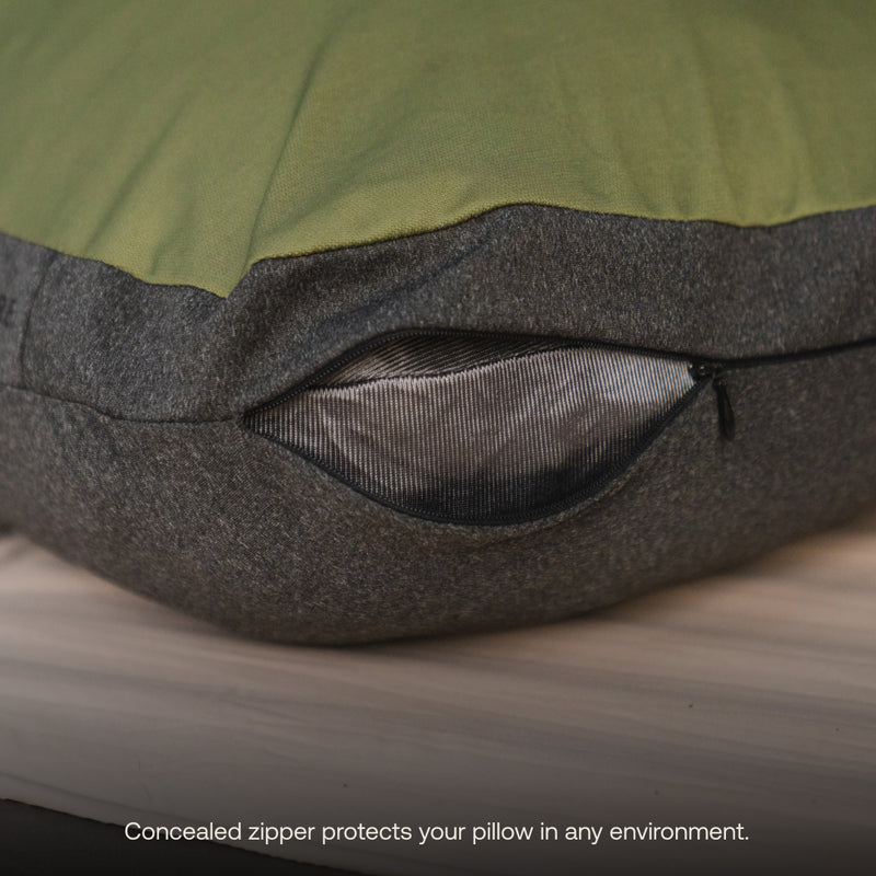 Close-up of a Luno® Camping Pillowcase showcasing a green top and gray bottom, with a partially opened concealed zipper revealing the cooling jersey knit fabric inside. Wooden surface in the background. Text overlay at the bottom reads, "Concealed zipper protects your pillow in any environment.