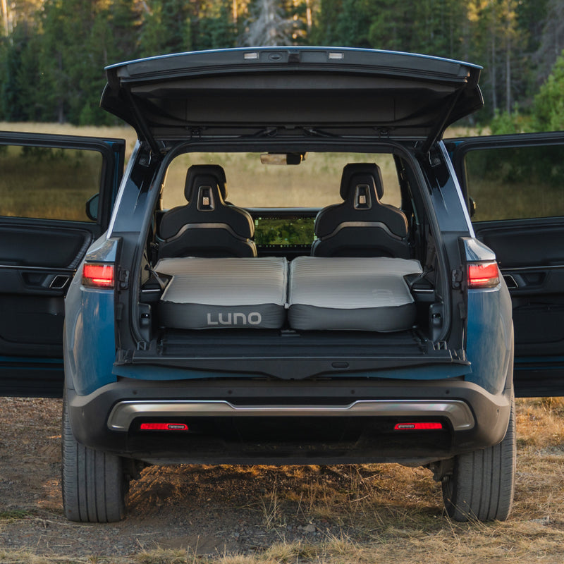 A blue SUV, likely a Rivian R1S, is parked with its rear hatch open, showcasing the innovative AIR+FOAM PRO Rivian R1S Mattress by Luno® inside the cargo area. This premium mattress offers a cozy setup for car camping within the spacious interior. Trees and greenery provide a serene background.