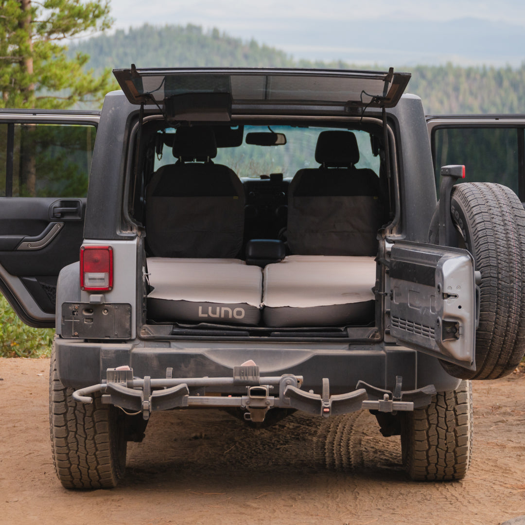 A Jeep with open rear doors showcases the back seat area converted into a cozy sleep system, featuring the Luno® AIR+FOAM PRO Jeep Wrangler Mattress. The vehicle is parked on a dirt path surrounded by trees, providing a scenic view that promises adventurous dreams.