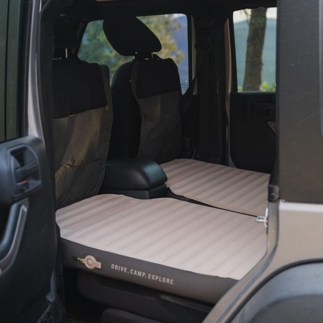 The image displays the interior of a car with its rear seats folded down, featuring a Luno® AIR+FOAM PRO Jeep Wrangler Mattress. The beige mattress fits perfectly within the vehicle, offering a flat sleeping surface ideal for adventurers.