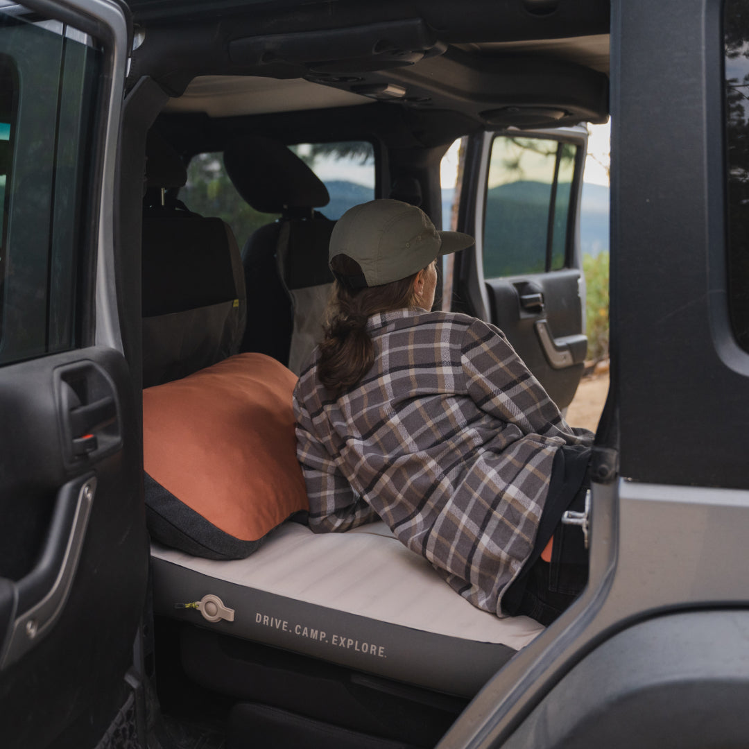 A person in a plaid shirt and cap is lounging in the back of a car equipped with the Luno® AIR+FOAM PRO Jeep Wrangler Mattress, enjoying a scenic view. The car door is open, displaying the words