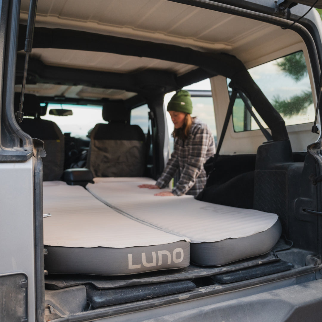 A person wearing a beanie and flannel shirt is setting up the AIR+FOAM PRO Jeep Wrangler Mattress from Luno® in the back of their vehicle, preparing for a cozy car camping experience with the Luno sleep system.