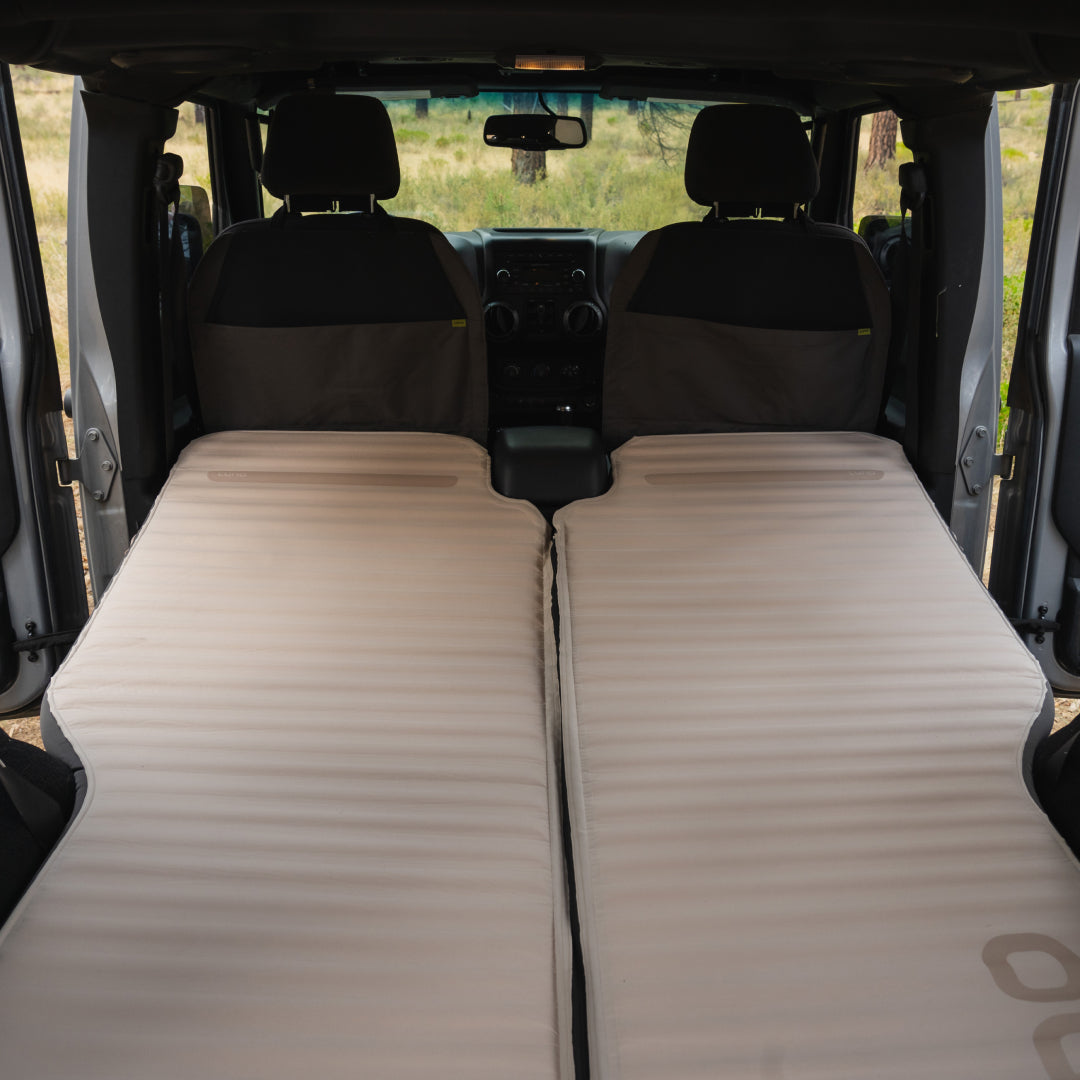 Inside the vehicle, the seats transform into a cozy sleep system with two beige AIR+FOAM PRO Jeep Wrangler Mattresses from Luno®. The mattresses ensure comfort as you gaze at the grassland through the windshield.