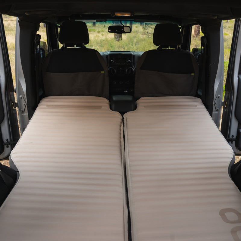 Inside the vehicle, the seats transform into a cozy sleep system with two beige AIR+FOAM PRO Jeep Wrangler Mattresses from Luno®. The mattresses ensure comfort as you gaze at the grassland through the windshield.