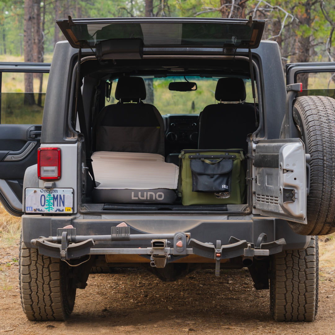 The rear of a parked Jeep Wrangler is shown with its back door open, revealing a Luno® AIR+FOAM PRO Jeep Wrangler Mattress inside. A green bag is also visible. The Jeep has an Oregon license plate and sits nestled in a wooded area.