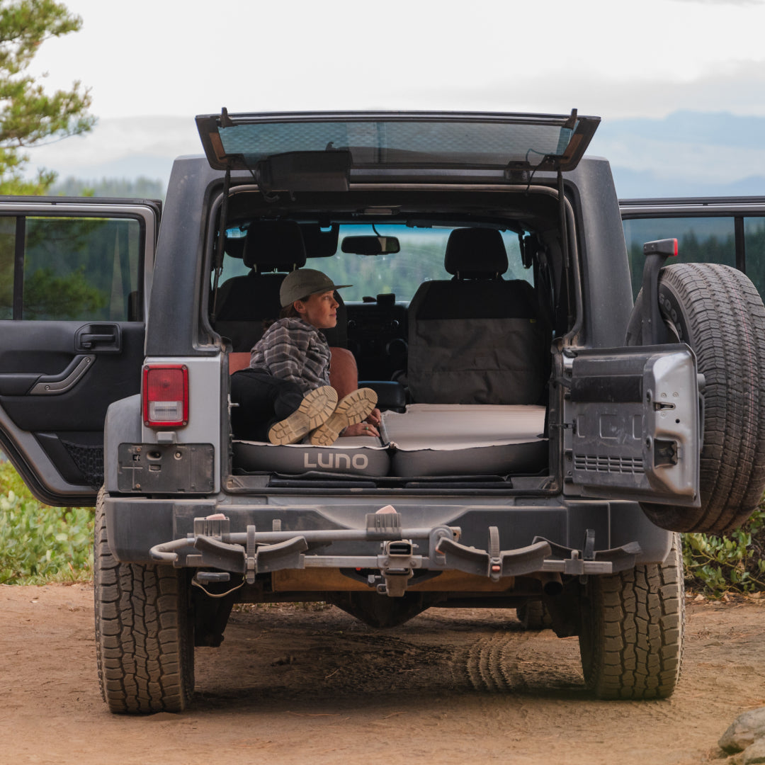 A person relaxes in the back of an SUV with the rear door open, surrounded by forest scenery. They're lying on a Luno® AIR+FOAM PRO Jeep Wrangler Mattress, enjoying the view. The vehicle is parked on a dirt path, with trees and mountains in the background.