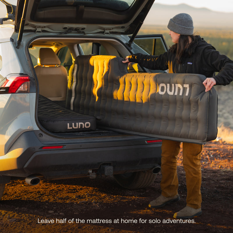 A person in a gray beanie and brown pants is unloading half of a Luno® AIR Pro Vehicle Mattress, featuring the "LUNO" logo, from an SUV's back. The setup suggests prepping for a solo camping trip with convenient solo zipper features.