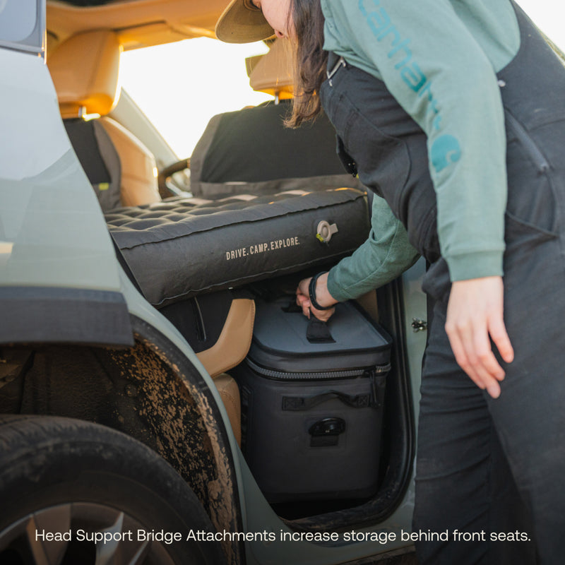 A person in outdoor clothing accesses a cooler in the back seat area of a vehicle with folded seats, covered by camping gear and an AIR Pro Vehicle Mattress by Luno®. Caption: "Head Support Bridge Attachments boost storage behind front seats.